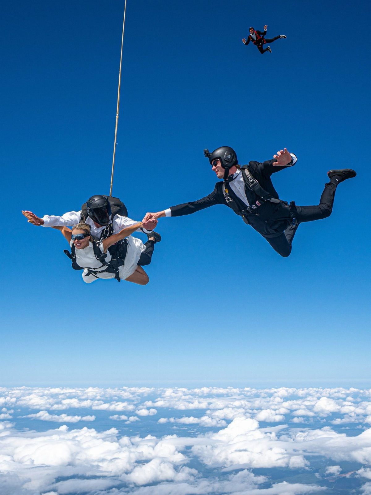 Skydiving couple in formal wear holding hands while falling through a bright blue sky