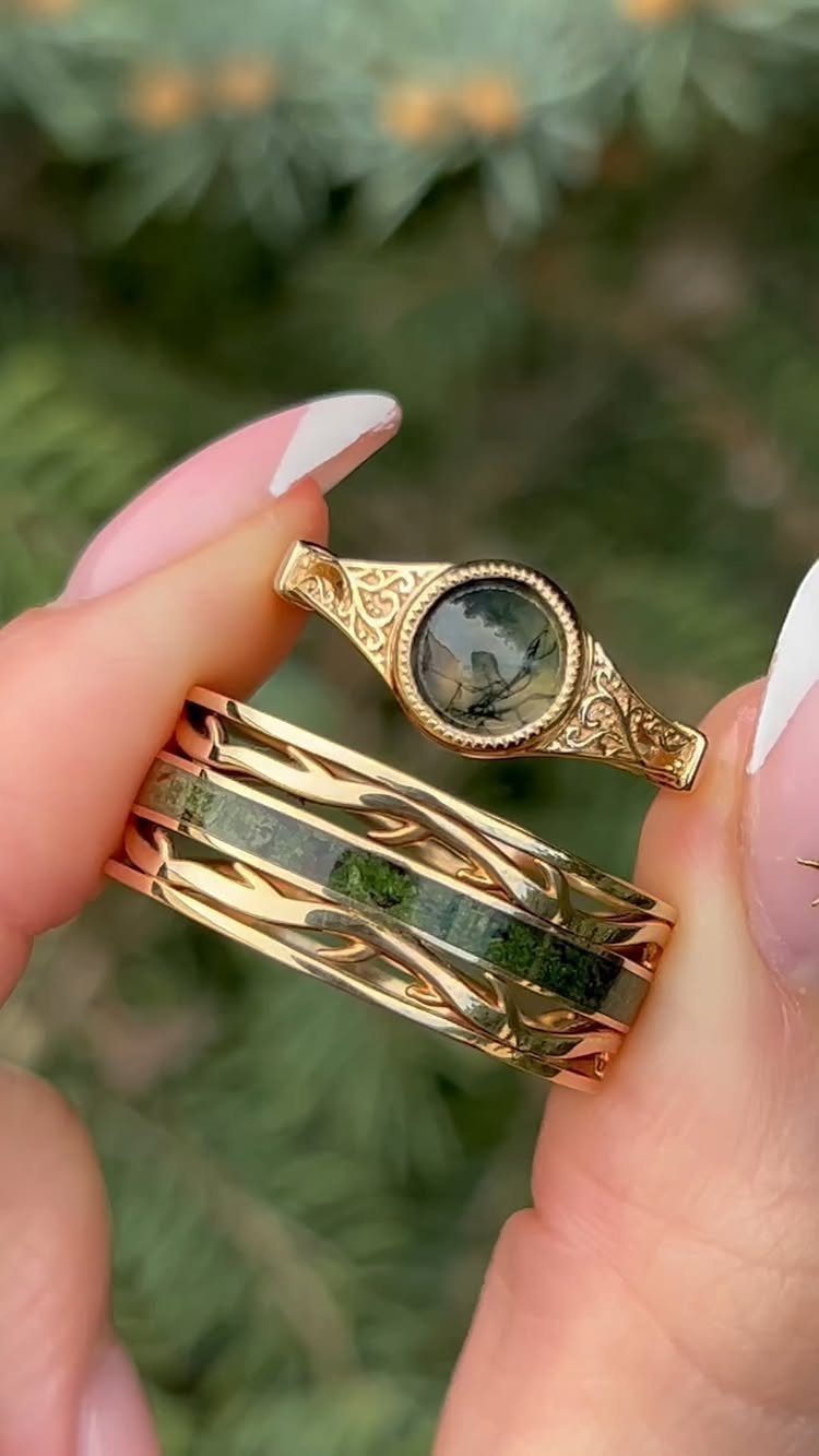 Two gold rings with leaf engravings and green mossy inlay held by fingers.