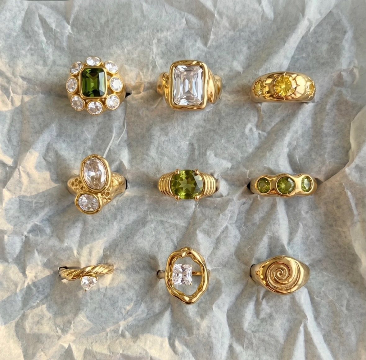 Nine gold rings with various gemstones arranged on crumpled grey paper.