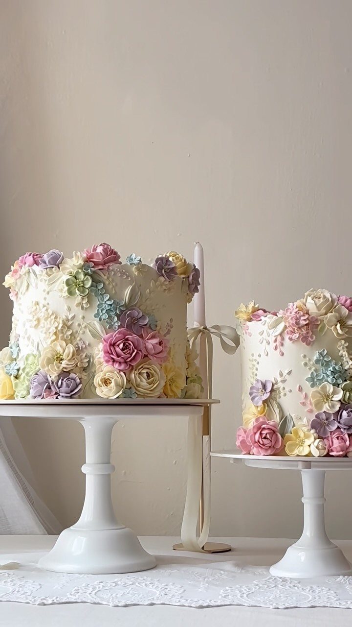 Two white cakes decorated with colorful buttercream flowers on white cake stands.