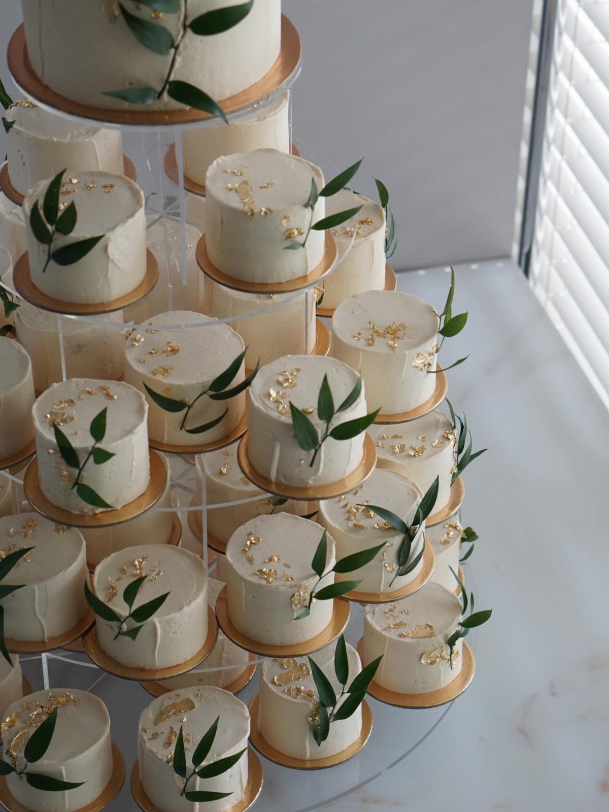 Mini white cakes with gold leaf and green leaves on a clear tower.