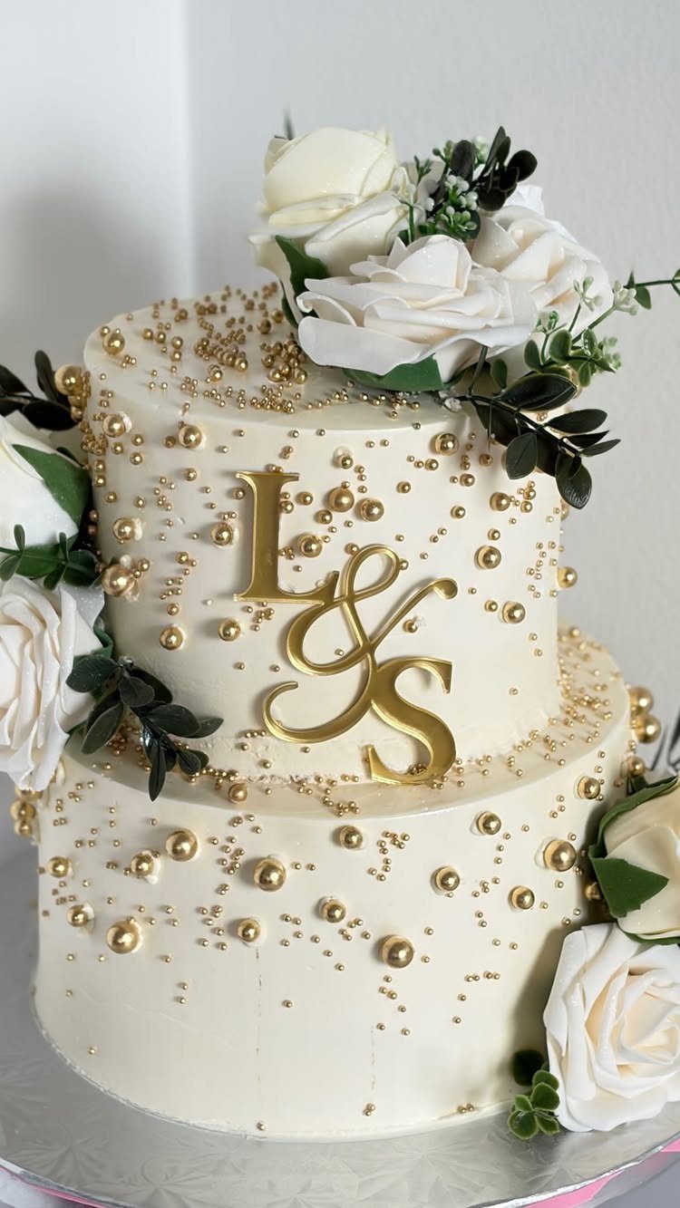 Two-tier white cake with gold monogram letters, gold pearls, and white roses.