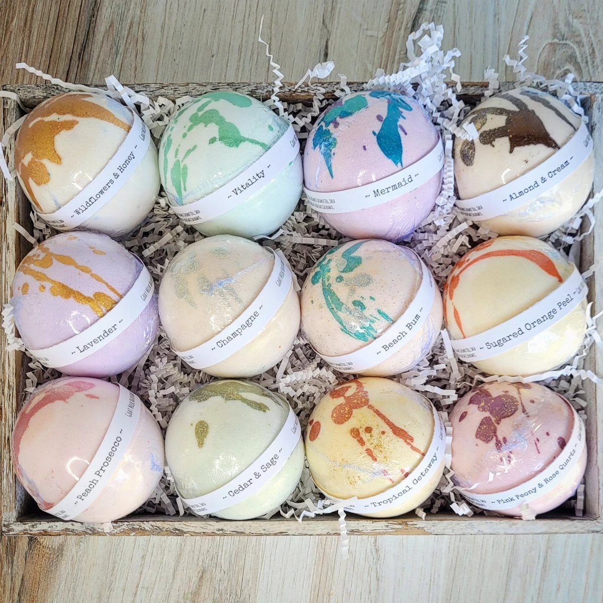 Twelve colorful bath bombs with labels arranged in a wooden crate with white filler.